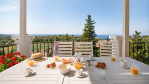 A white table set with breakfast items, including orange juice, eggs, and pastries, on a balcony with a scenic view. - Villa Katerini in Paxi (Paxos)