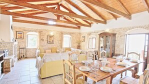 A rustic room with a stone fireplace, a wooden dining table set for four, and a bed with a yellow headboard. - Villa Katerini in Paxi (Paxos)