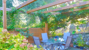 A covered patio area with a table and chairs, surrounded by greenery and a wooden fence.