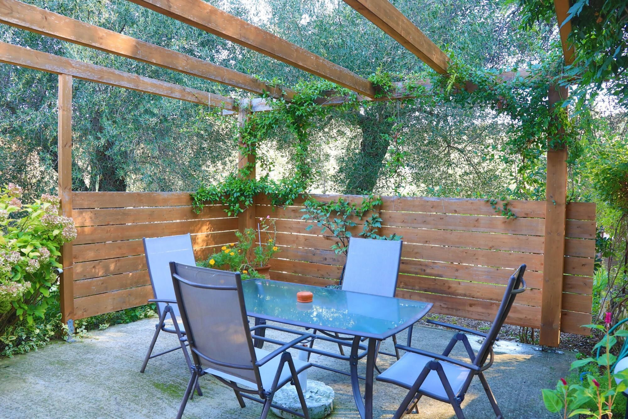 A wooden pergola with a glass table and metal chairs in a garden area.
