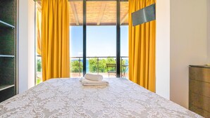Ein Schlafzimmer mit einem großen Bett, gelben Vorhängen und Blick auf Grünflächen sowie ein Gewässer.
