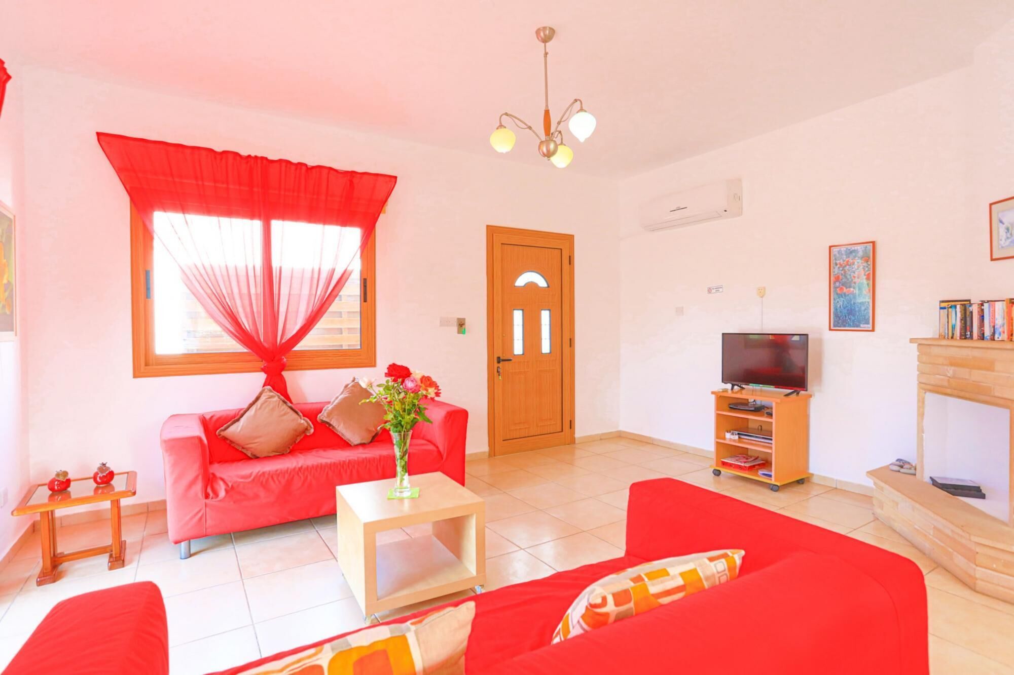 A living room with a red sofa, a TV stand with a flat-screen TV, a wooden door, and a window with sheer curtains.