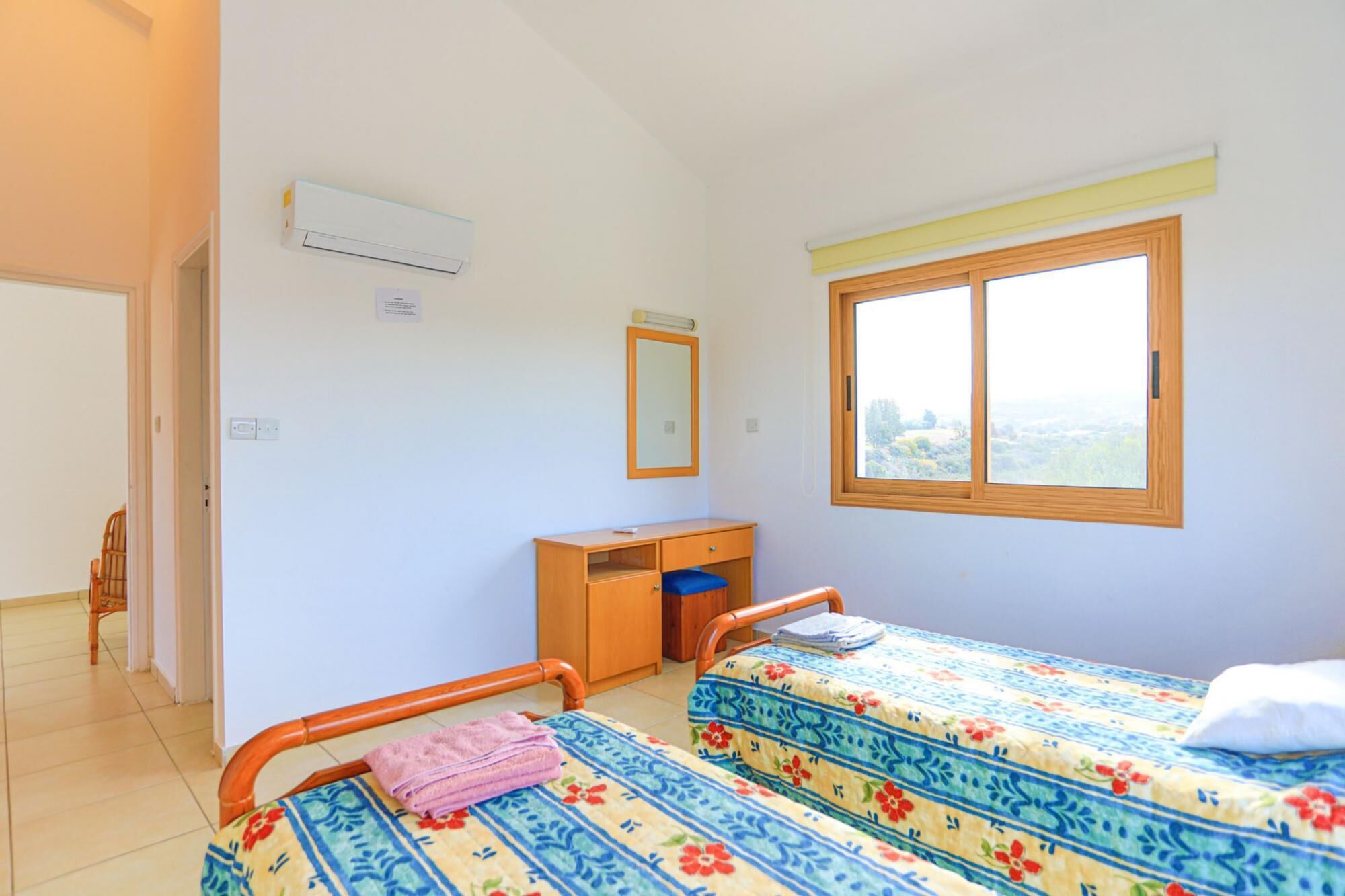 A room with two single beds, a wooden desk, a mirror, and a window with a view.