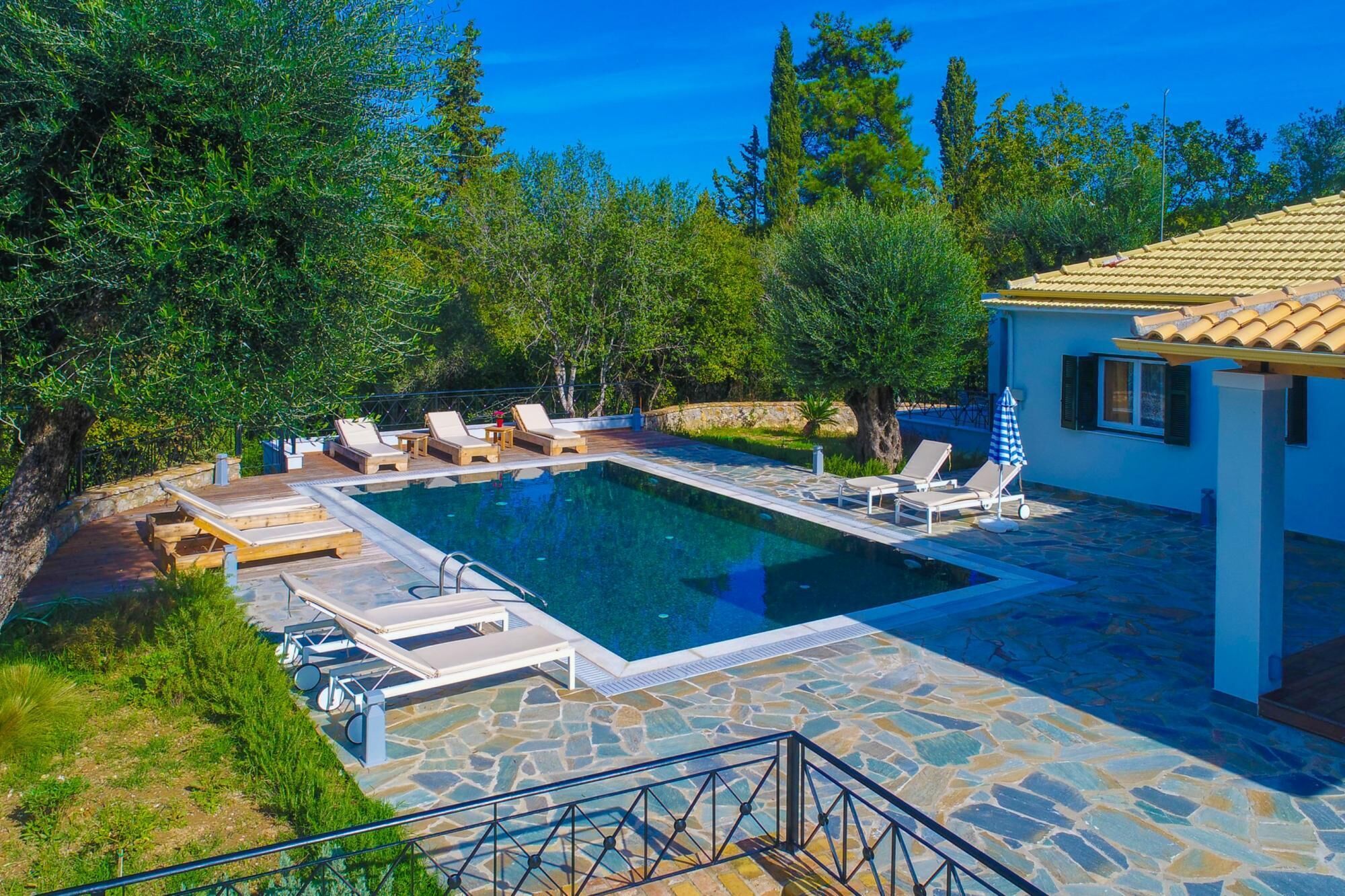 Une piscine rectangulaire entourée d’une terrasse en pierre, de chaises longues et d’un petit bâtiment doté d’un toit recouvert de tuiles.