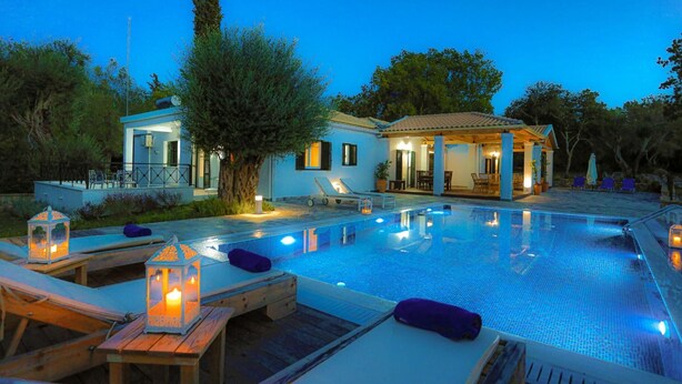 A swimming pool with lounge chairs and a covered seating area, surrounded by trees and a building with a tiled roof. - Achilleion Forest Villa (Corfu)