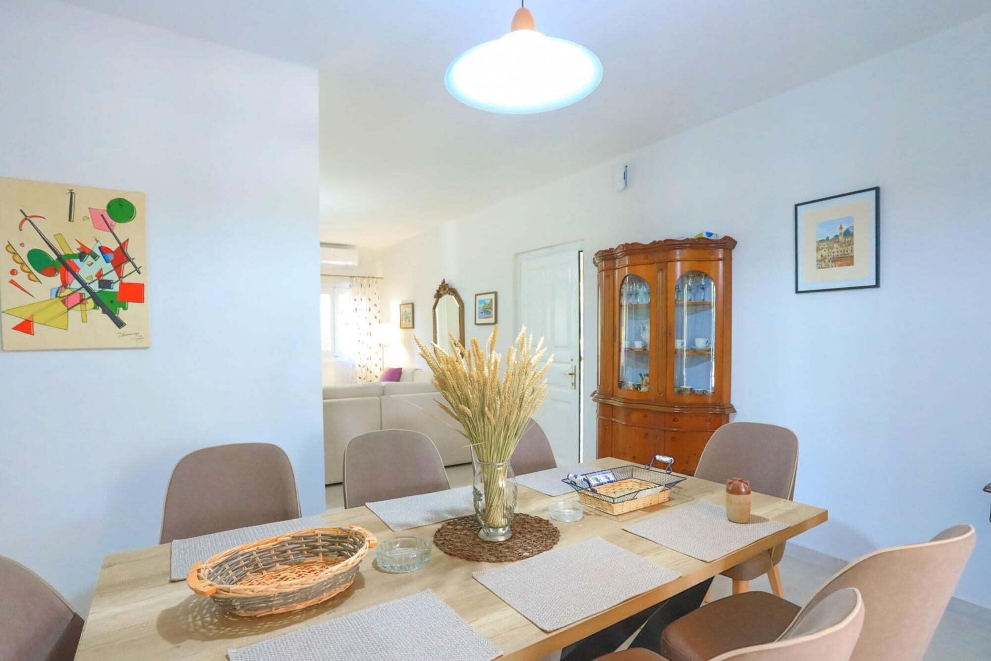 A dining area with a wooden table, chairs, a vase with dried plants, a cabinet, and a painting on the wall.