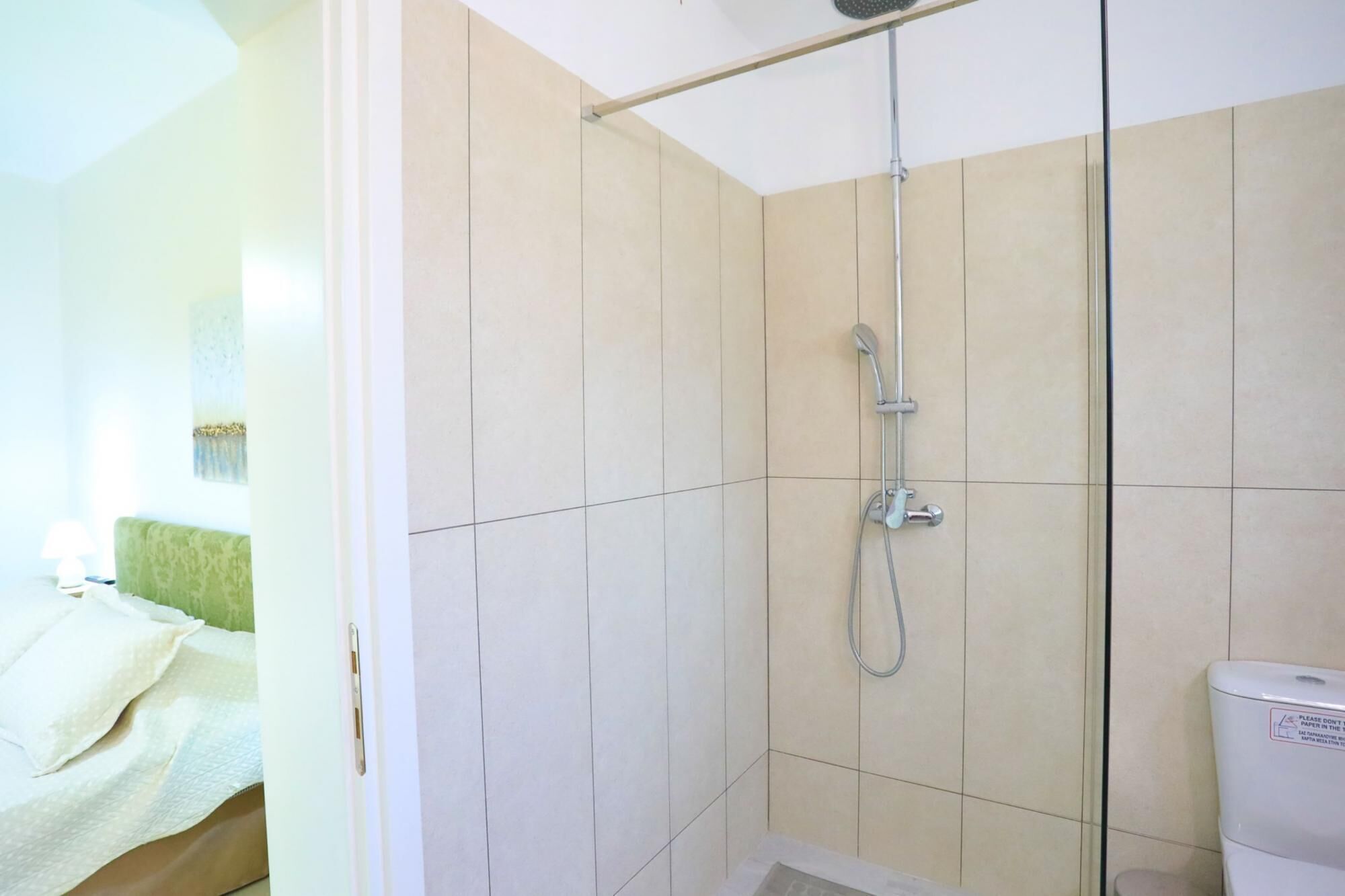 A shower with a handheld showerhead, a glass-enclosed toilet, and a bed with a green headboard.