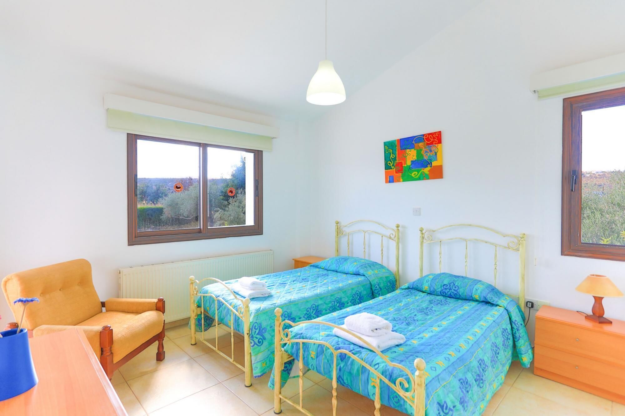 A bedroom with two single beds, a chair, a table, a radiator, a window with a view, and a colorful artwork on the wall.