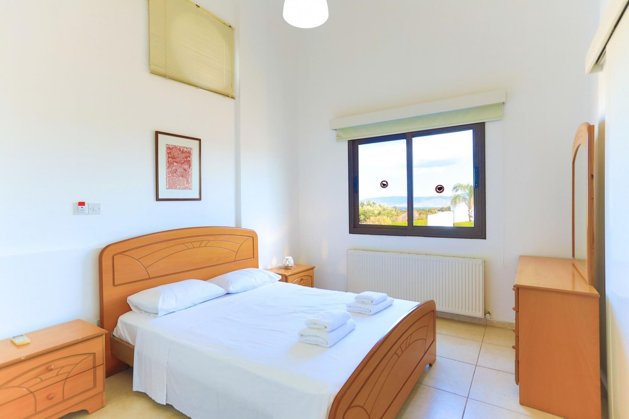 Ein Schlafzimmer mit einem hölzernen Bett, einem Nachttisch, einer Kommode, einem Heizkörper und einem Fenster mit Blick ins Freie.