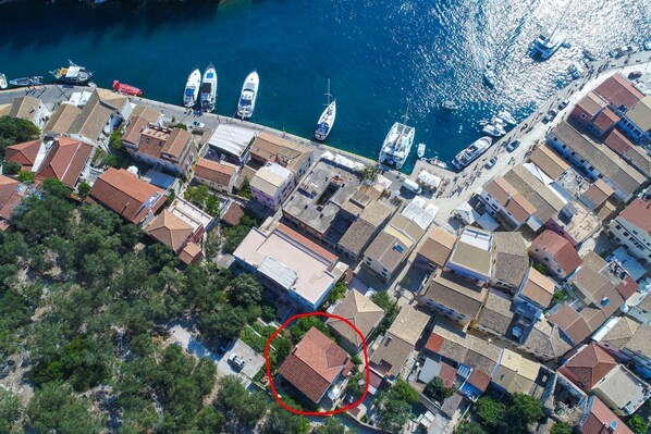 An aerial view of a coastal town with terracotta-roofed buildings, a marina with several boats, and a circled building with a distinct triangular roof. - Villa Eleni Paxos in Gaios (Paxos)