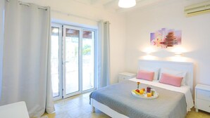 A bedroom with a bed, a bedside table, a nightstand, a window with curtains, and a wall-mounted air conditioning unit. - Paola House in (Corfu)