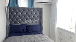 A bedroom with a tufted headboard, a grey and blue bedspread, and a white dresser with drawers.