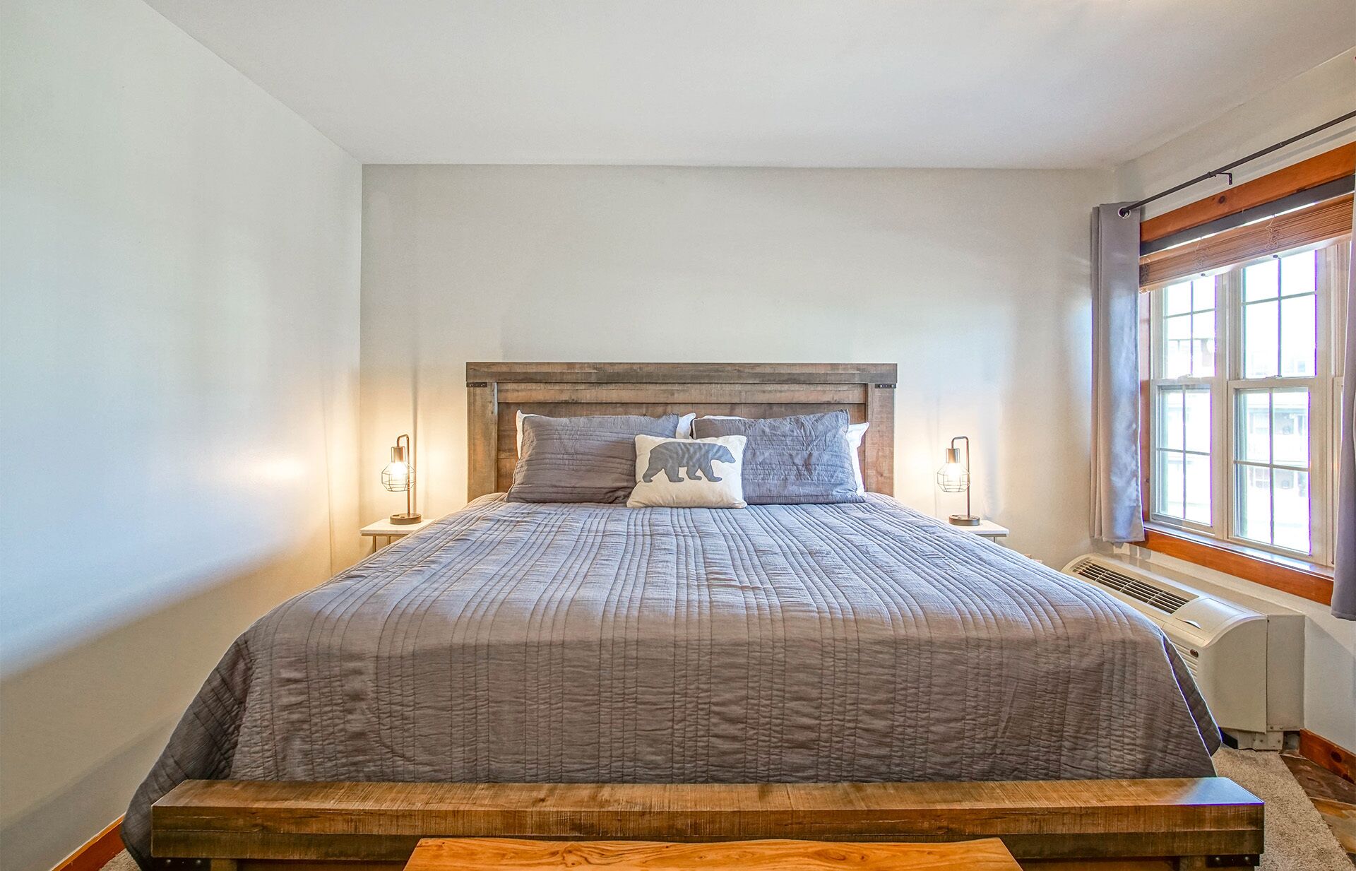 Ein Schlafzimmer mit hölzernem Kopfteil, einem Bett mit grauer Steppdecke, zwei Nachttischlampen und einem Fenster mit Vorhängen.