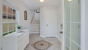 Um corredor com escada, uma porta branca, uma janela com persianas e um vaso com planta.