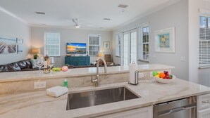 Uma cozinha moderna com bancada de mármore, pia de aço inoxidável e lava-louças. Há uma área de refeições com um sofá de couro marrom e uma TV exibindo uma cena de pôr do sol.