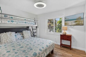 Un dormitorio con una cama individual sobre otra, una mesita de noche con lámpara, una ventana con persianas y un cuadro con motivos playeros en la pared.