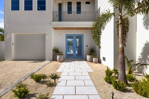 A modern two-story house with a blue door, a garage, and a gravel driveway with potted plants. - Beach Walk: Ultimate Luxury! Games Galore, Pool, Spa Rooftop Deck (Holmes Beach)