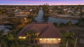 Una casa con techo rojo, rodeada por un pequeño lago, palmeras y un camino de acceso bien iluminado.