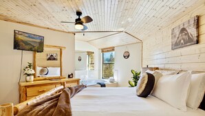 Ett sovrum med ett träpanelat tak, en säng med vita sängkläder, en byrå i trä, ett skrivbord, en stol och ett stort fönster med utsikt över träd.