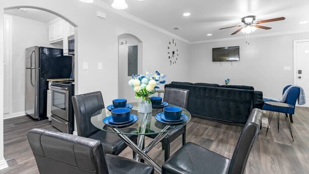 A modern kitchen with stainless steel appliances, a dining area with a glass table and blue chairs, and a living area with a dark sofa and a ceiling fan.