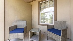 A small outdoor seating area with two wicker chairs and a glass table, positioned against a wall with a window.
