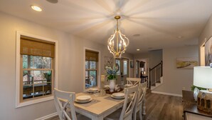 A dining area with a white table set for four, surrounded by white chairs, a chandelier, and a view of the outdoors through large windows with blinds.