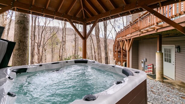 A hot tub under a wooden pergola with a view of a forested area and a building with a balcony.