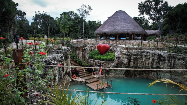 Edifici amb teulada de palla envoltat de vegetació exuberanta i una piscina de color blau clar.