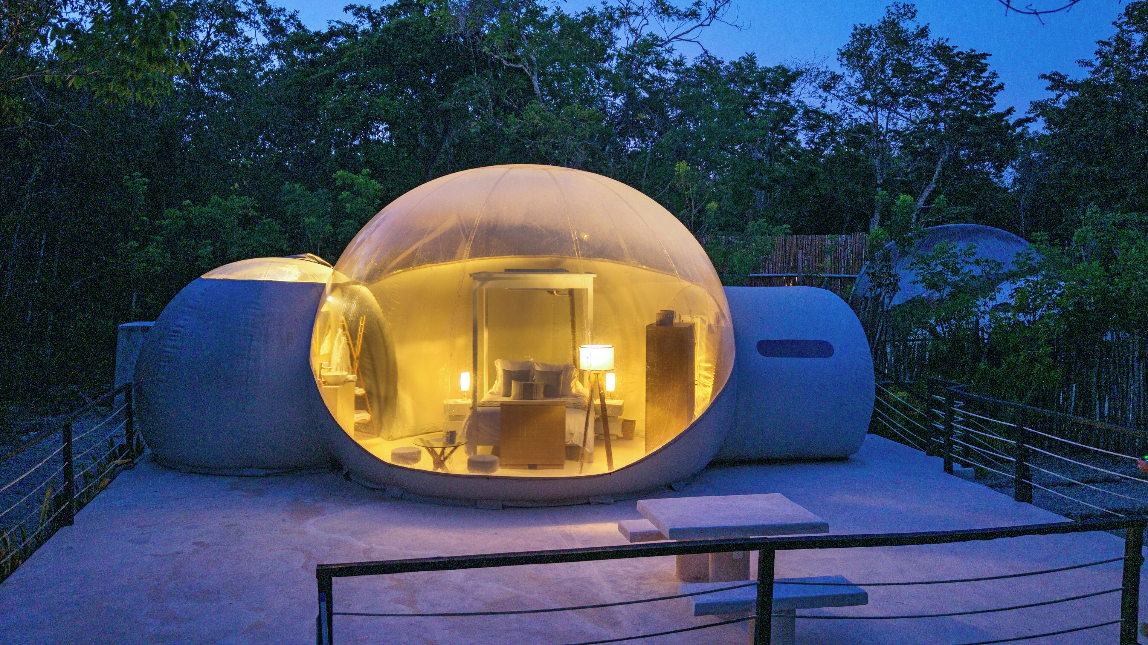 A spherical, illuminated structure with a bed and a desk, surrounded by trees.