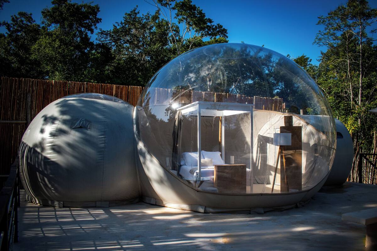 Estructura esférica con una sección transparente que muestra una cama y una mesita, rodeada por una valla de madera y árboles.