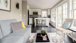 Ein modernes Wohnzimmer mit einem grauen Sofa, einem Couchtisch mit Tablett und einem Sessel. Im Hintergrund ist eine Küche mit weißen Schränken und einem Essbereich zu sehen.