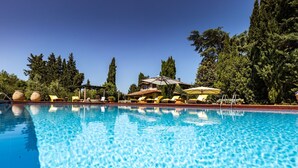Une piscine aux eaux cristallines, bordée de chaises longues et d’ombrelles, entourée d’arbres et avec un bâtiment visible au loin.