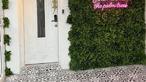 Una porta bianca con un pannello in vetro, una maniglia a forma di coccodrillo dorato e una piccola finestra. Un neon recita: “Find me under the palm trees.”