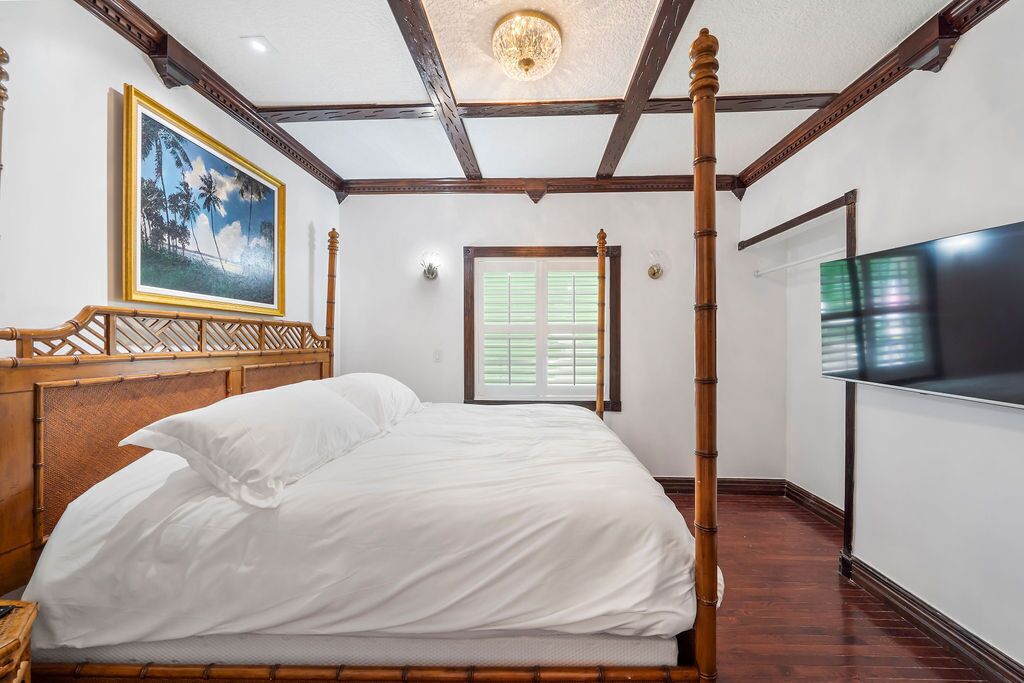 A four-poster bed with white linens, a wooden headboard, and a painting of a tropical scene on the wall.