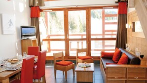 Uma sala de estar moderna com uma mesa de jantar de madeira, cadeiras vermelhas, um sofá com almofadas laranja e uma televisão sobre um suporte.