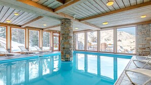 En inomhuspool med pelare av sten, träbjälkar och stora fönster som ger utsikt över ett snötäckt landskap.