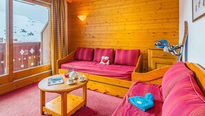 Ett mysigt rum med en väggpanel av trä, en soffa med röda kuddar, ett litet bord med en tekanna och koppar, och ett fönster med utsikt över ett snöklätt berg.
