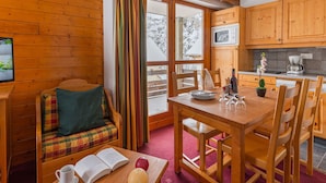 Een knus interieur van een chalet met een houten eettafel, stoelen, een bank met kussens, een raam met gordijnen en een keuken met houten kasten en een magnetron.