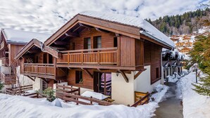 A wooden chalet with a balcony, a snow-covered roof, and a path leading to a village. - Lovely apartment for 6 people with WIFI, pool, TV, terrace and pets allowed (Les Gets)