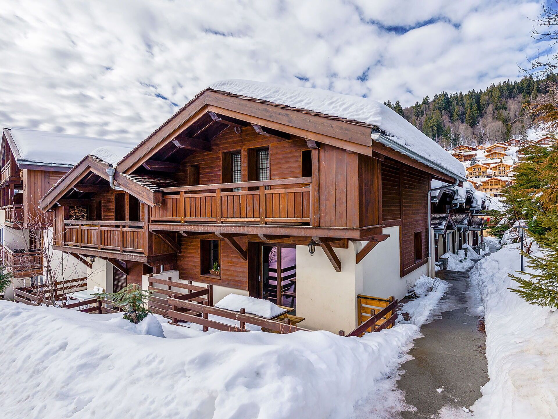 A wooden chalet with a balcony, a snow-covered roof, and a path leading to a village.