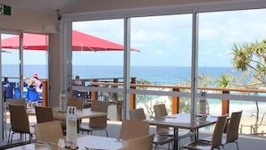 An outdoor dining area with tables and chairs, large windows with a view of the ocean, and red umbrellas. - Top 1 Sunshine Beach Family Retreat (Sunshine Beach)