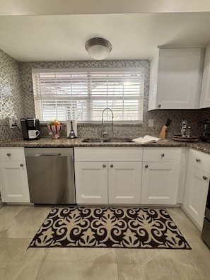 A kitchen with a dishwasher, a sink, a window with blinds, and a patterned rug.