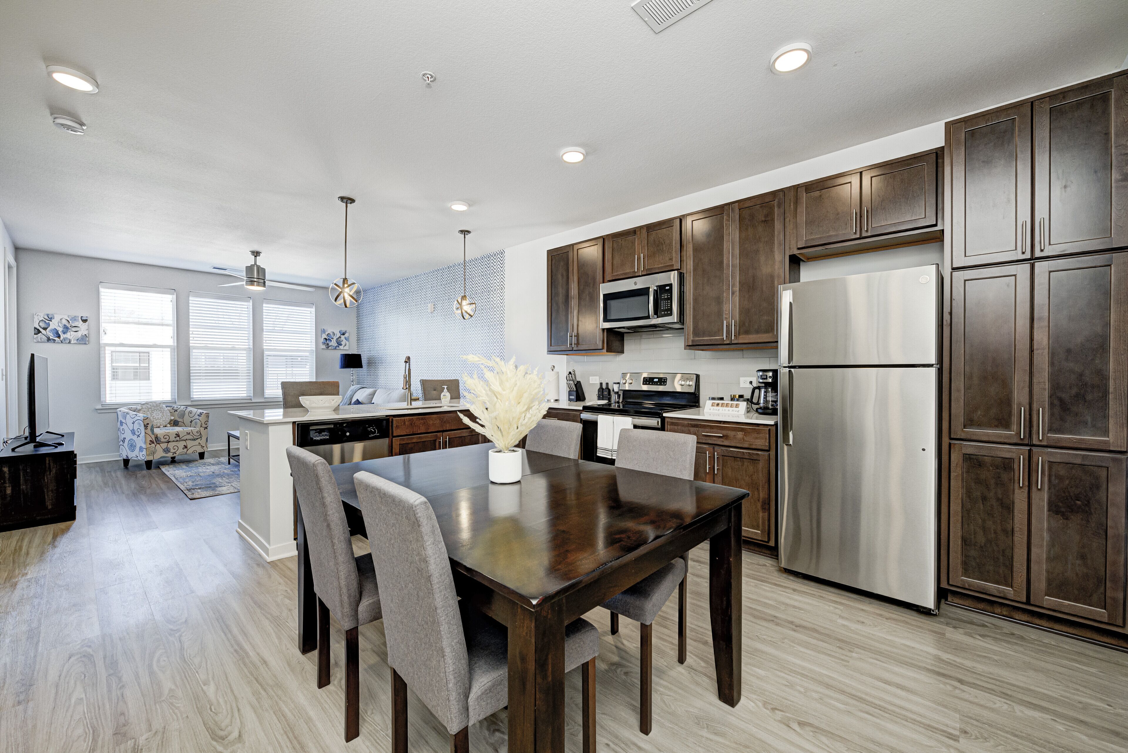 A modern kitchen with dark wooden cabinets, stainless steel appliances, and a dining area with a dark wooden table and grey chairs.