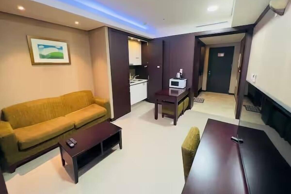 Un espacio habitable pequeño con sofá, una mesita, kitchenette y una puerta que da a otra habitación.