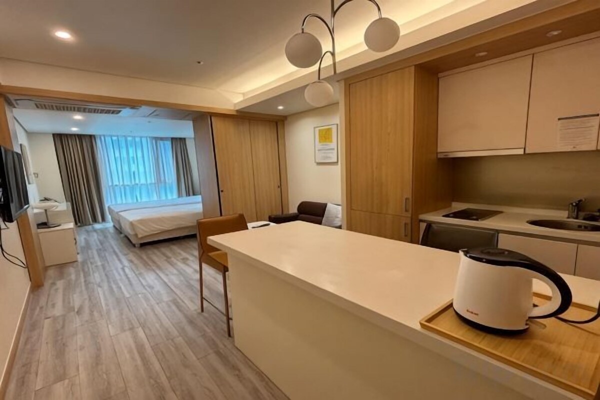 A modern hotel room with a kitchenette, a bed, a seating area, and a view of the outdoors.