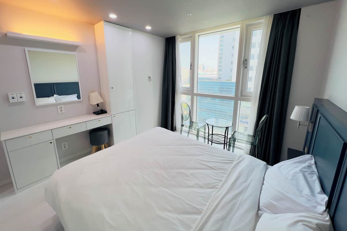 Habitación de hotel con una cama grande, un escritorio con lámpara, un espejo y vistas a la ciudad a través de amplias ventanas.