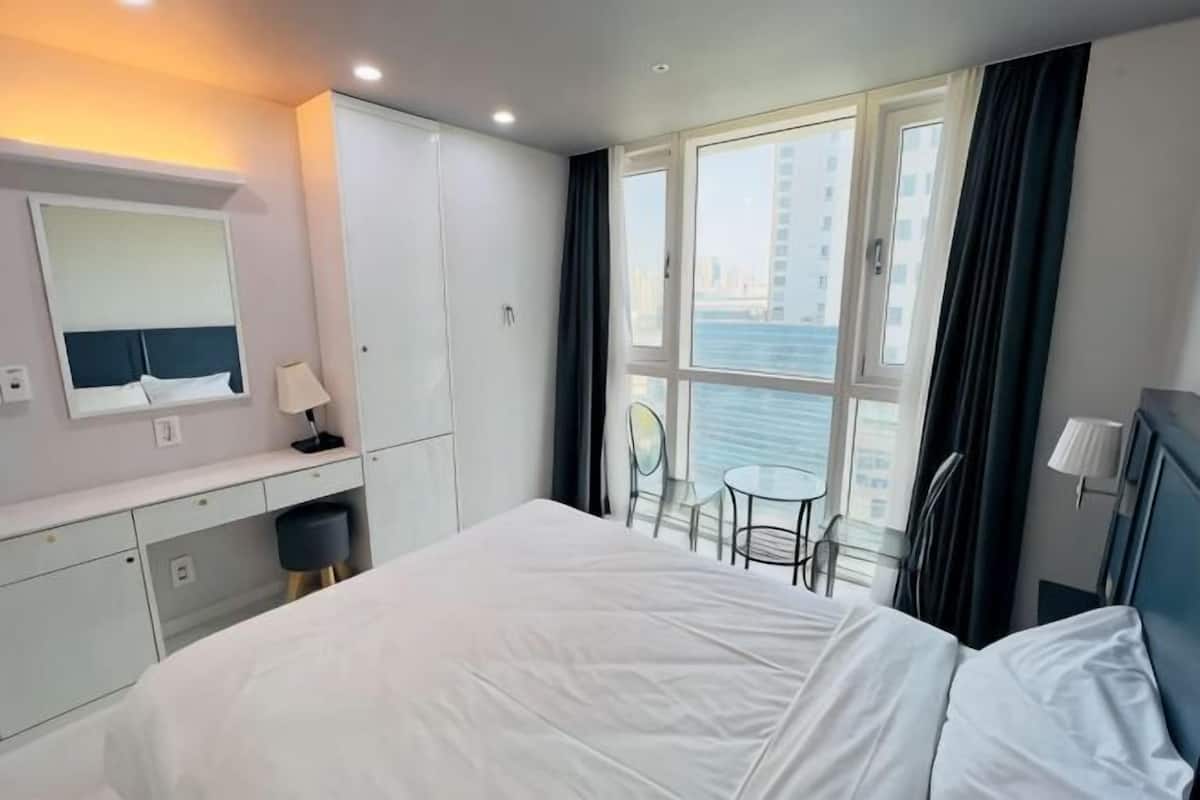 Habitación de hotel con cama, escritorio, silla, mesita, espejo y ventana con vista al agua.