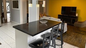 A modern kitchen with a central island, bar stools, a dining area with a sofa, and a TV in a yellow-walled living room. - Spacious 4 Bdrm House in Brampton (Brampton)