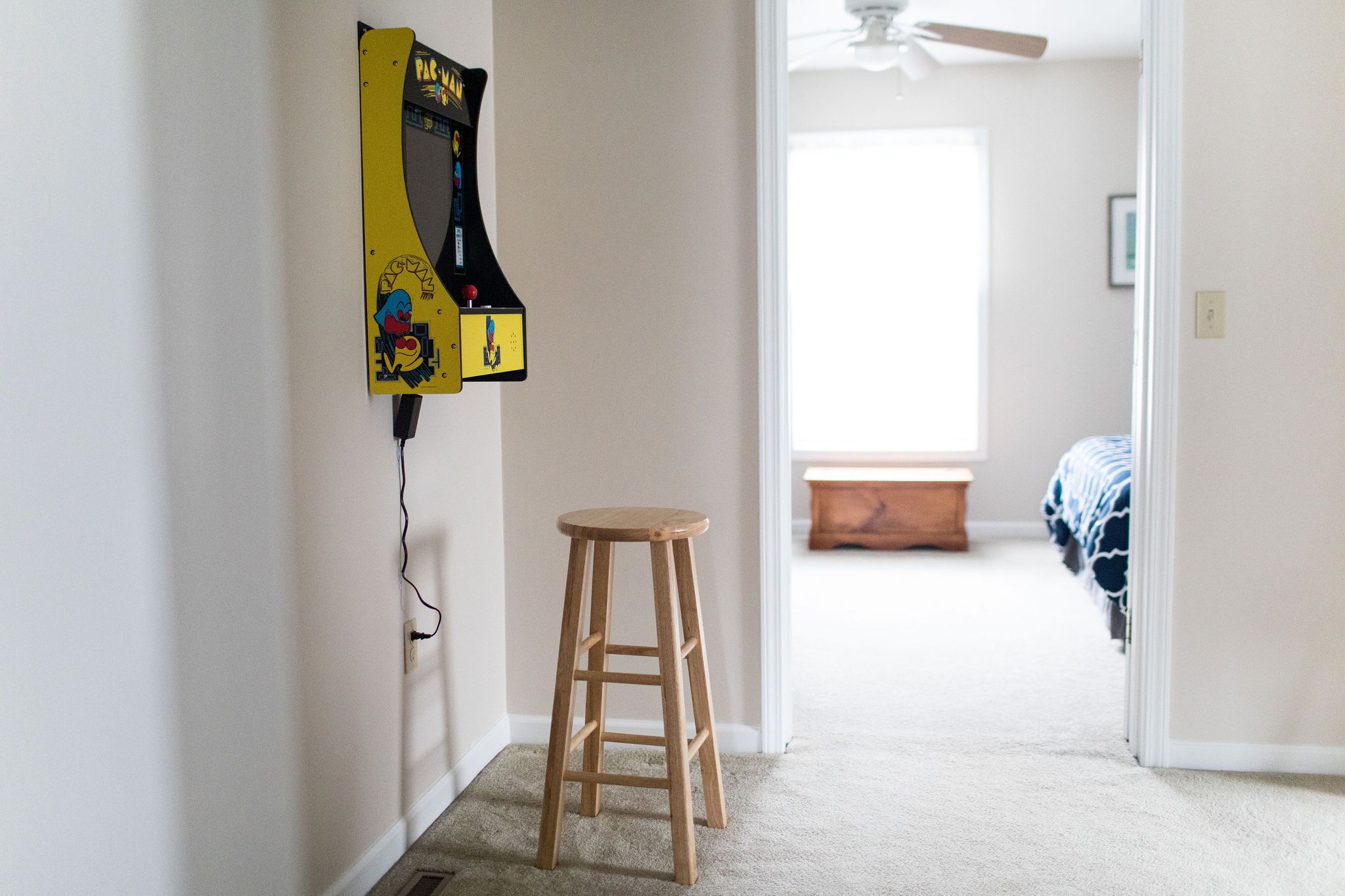 Un couloir avec un tabouret en bois, une borne d’arcade fixée au mur et un lit à l’arrière-plan.