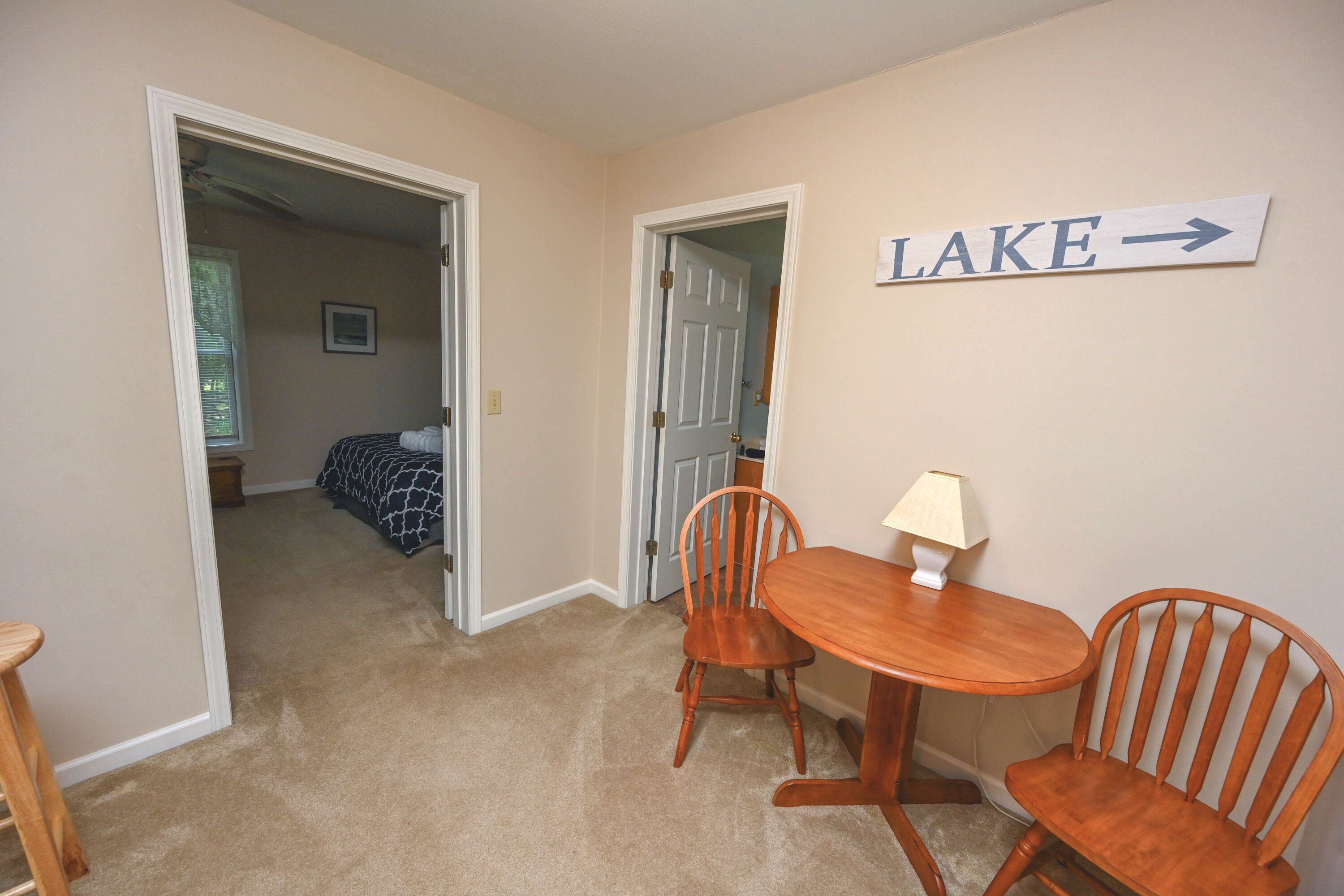 A room with a dining table and chairs, a sign pointing to the right labeled "LAKE", and an open door leading to another room with a bed and a window.